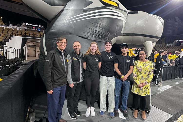 A group shot of six people, from left are two men, then a woman and two young men. A woman in a yellow top is on the right. They are standing in front of a large inflatable mascot.