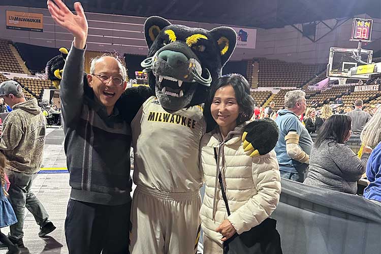 Man and a woman pose with a Panther mascot. The man on the left is wearing wire glasses and is waving at the camera. The woman on the right is wearing a cream-colored jacket and clutching a handbag.