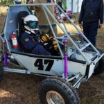 A driver poses in a white Baja car with a number 47 in dark blue on the side.