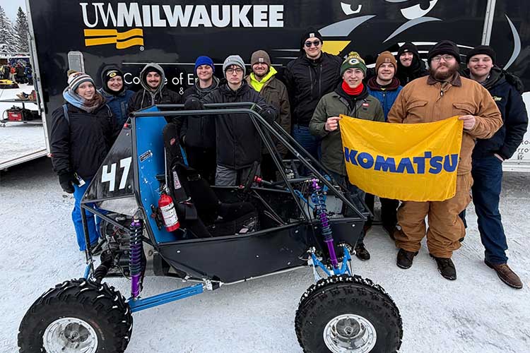 Twelve students pose with a blue and black Baja car they built. Two are holding a yellow flag with "Komatsu" in blue letters.