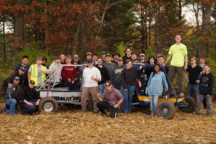 A group shot of 20 students posed around two Baja cars, looking at the camera.
