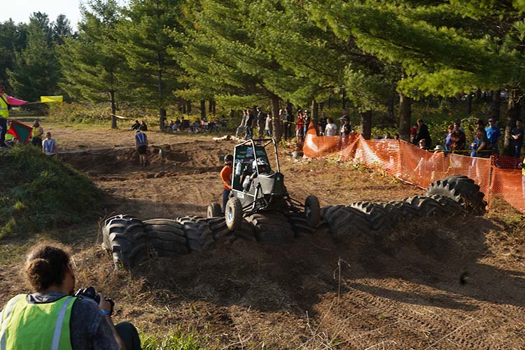 an all-terrain Baja car gets over a bank of tires. The back of one person's head and sholders is visible in the far left bottom of image.