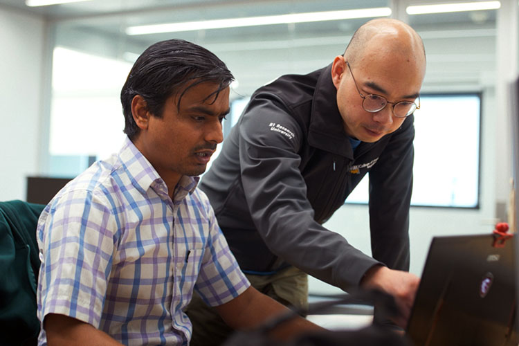 Two men staring at a computer screen. The one on the left wearing a white, short-sleeved shirt and has dark hair. The man on the right has glasses, is wearing a black vest and long-sleeved t-shirt, and is bald.