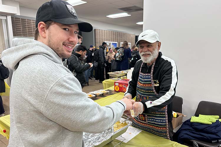 A bearded man with white cap and apron on right serves up soup for a student in a light gray hoodie and dark baseball cap.