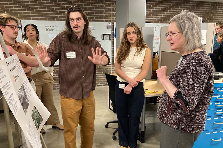 Two young men and two young women stand near a research poster on the left. The man in the center, with shoulder-length hair and a brown shirt on, gestures as he explains the work on the poster to a middle-age woman (far right)