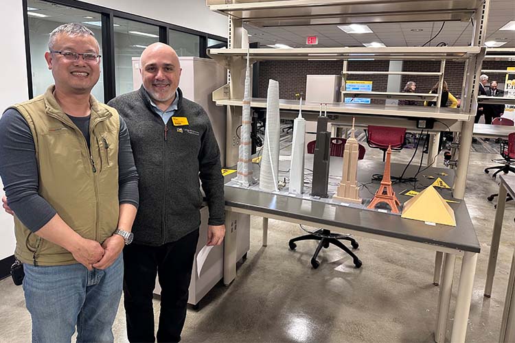 Two smiling men on the left look at the camera next to a display of 3D printed miniature buildings arranged by height (right).