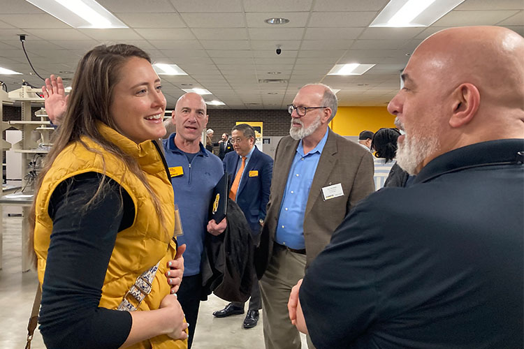A woman in a gold puffer vest (left) is conversing with a bald man in a navy polo shirt (right). Two men are in between and back, having their own discussion. Both are wearing blue shirts.