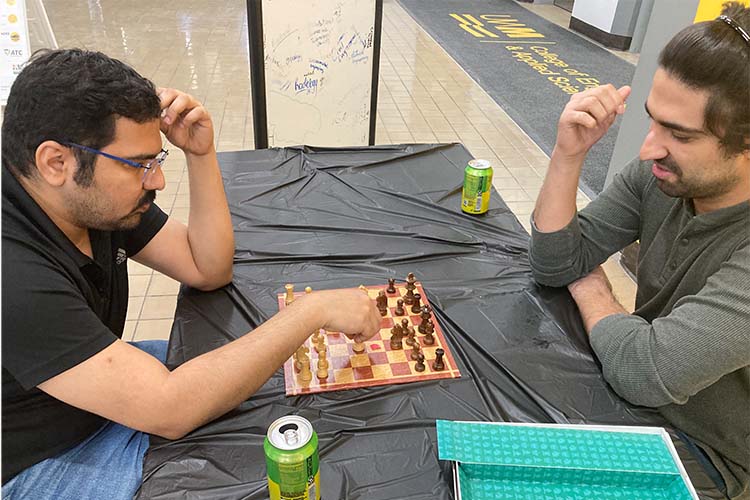 Two men play chess. The one on the left has glasses and a black T-shirt on, while the other is wearing a gray long-sleeved shirt.