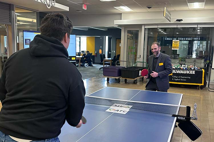 Two men are playing ping-pong. The one of the left is seen from the back. A bearded opponent is serving.