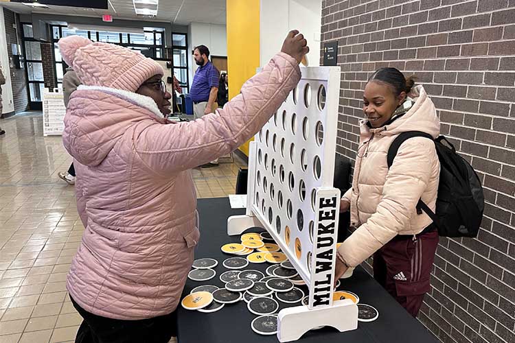 Two young black women are playing a game of Giant Connect Four. The one on the left is wearing a pink coat and hat. The other has on a cream-colored coat.