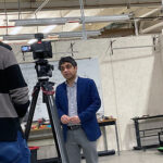 Two men, one with glasses with his back showing, are talking to each other with a camera on a stand in between them. The man seen from the front is wearing a blue shirt and navy jacket.