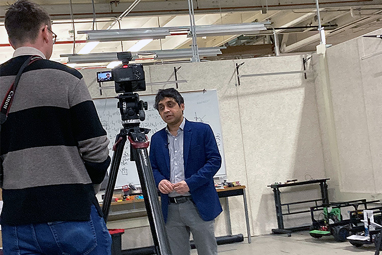 Two men, one with glasses with his back showing, are talking to each other with a camera on a stand in between them. The man seen from the front is wearing a blue shirt and navy jacket.