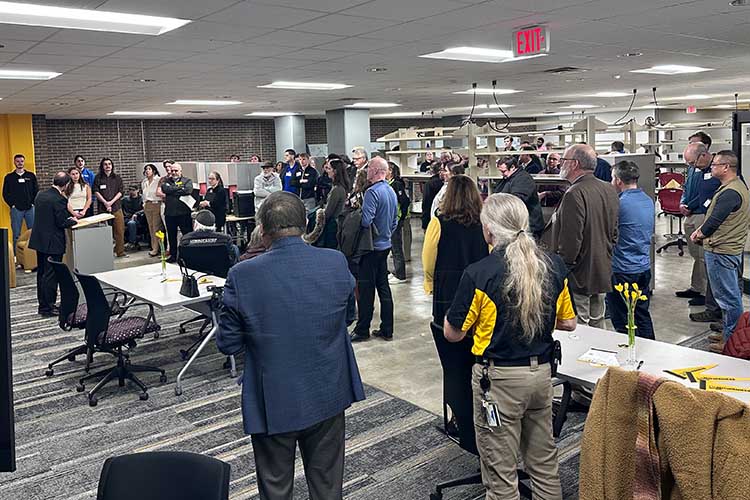 A crowd of about 30 people face a person standing at a podium (left) at a gathering.