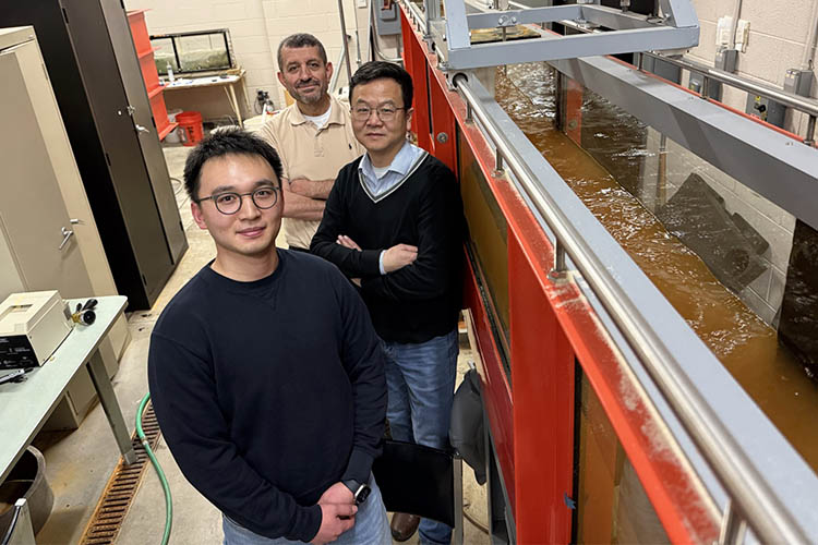 Three stand front to back looking at the camera and standing next to a large tank of moving water. The man in front is wearing a navy sweatshirt; the one in the middle wears a navy sweater and has glasses; and the one in the back is wearing a light tan polo shirt.
