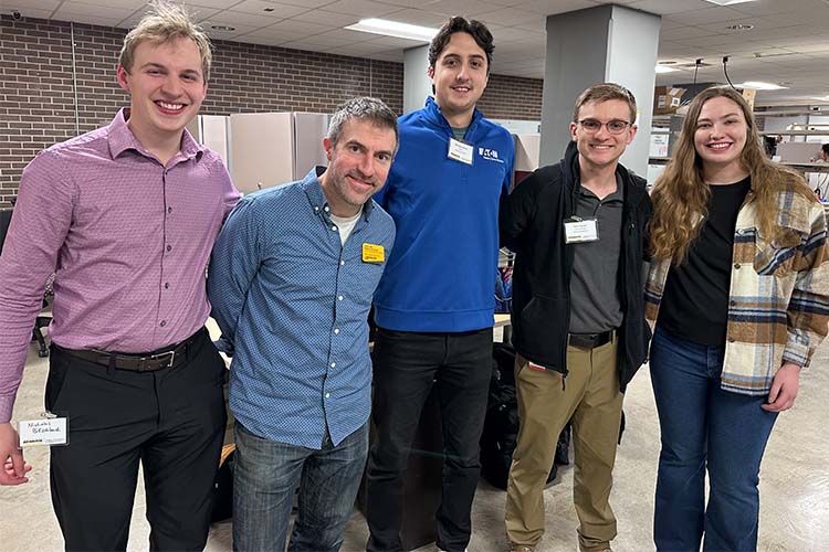 Five people, including one woman, pose for the camera, arm in arm. The man on the far left is wearing a lilac shirt, the next two are in blue, the fourth is wearing a dark gray shirt and jacket, and the woman has on a brown and beige plain shacket.