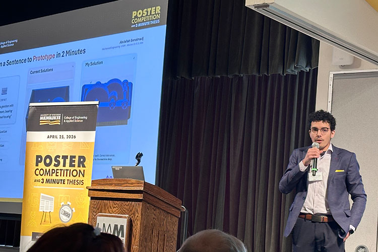 A young man with dark hair in a dark blue jacket and white shirt speaks while holding a microphone at an engineering and computer science research competition. A large screen and a banner is to his right