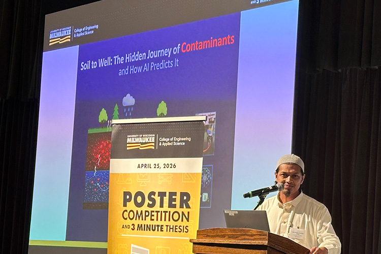 A man stands at a podium. He is smiling and wearing a long white tunic, traditional dress in Bangladesh, and cap.
