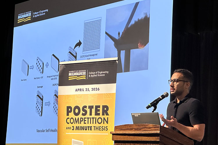 A man with very short dark hair and glasses speaks are a podium. Behind him is a large screen and to his right is a yellow banner.