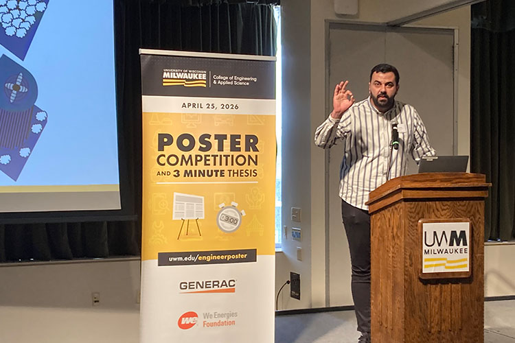 A man with dark, short hair and a white shirt with black stripes make a hand gesture while speaking at a podium. A yellow banner is to his right.
