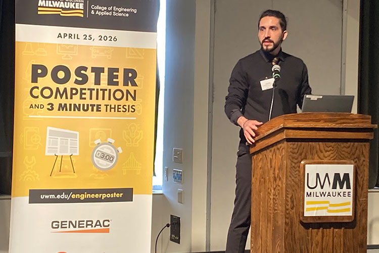 A man with dark hair pulled into a man-bun with black clothing addresses an audience at a podium. A banner is to his right.