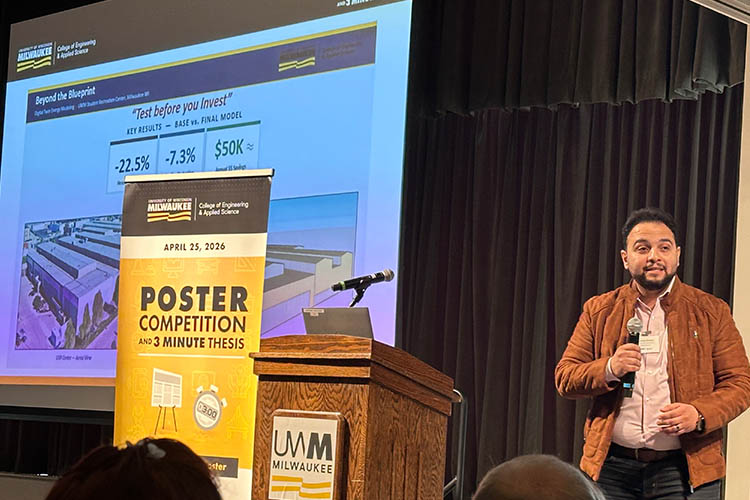 A man with dark hair and brown jacket speaks from a stage, microphone in hand. A large screen and banner are at his right.