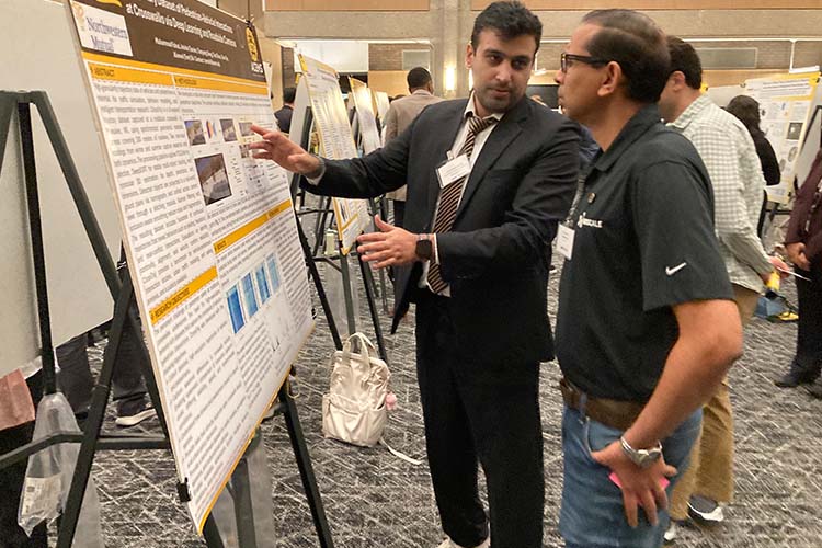 Two men standing side by side are looking at an engineering research poster.