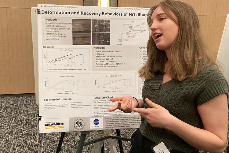 A young woman with sandy hair gestures as she explains her research poster to someone off-camera.