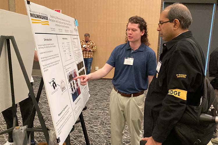 A younger man in a navy shirt (left) points out a fact on his engineering research poster to an professor/judge.