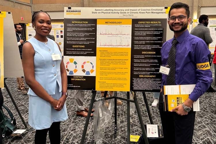 A Black woman in a light blue suit, left, displays here engineering research posters to a judge, right. The poster is in the middle.