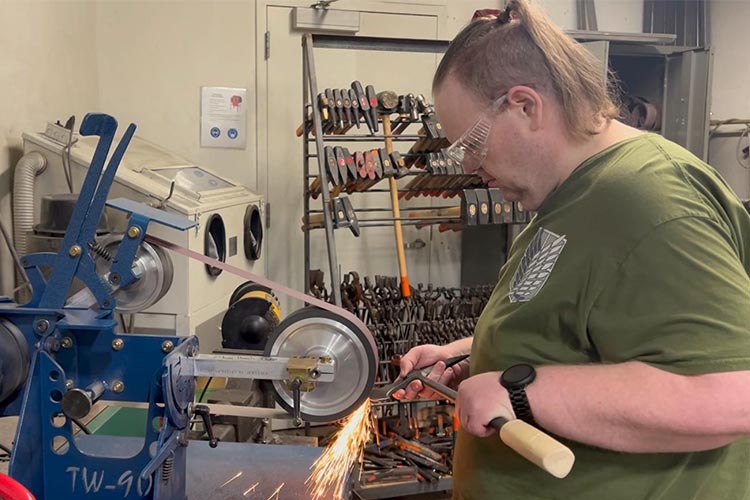 A man wearing an olive T-shirt with his hair in ponytails sharpen a blade with sparks flying.