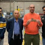 Four men looking at the camera. All are wearing dark colors except the third from the left who is in an orange sweatshirt and holding a metal alloy that he cast.