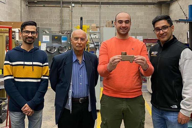 Four men looking at the camera. All are wearing dark colors except the third from the left who is in an orange sweatshirt and holding a metal alloy that he cast.