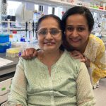 Two women from India look at the camera and smile. The younger one is standing behind the older one, with her hands on the older's shoulders.