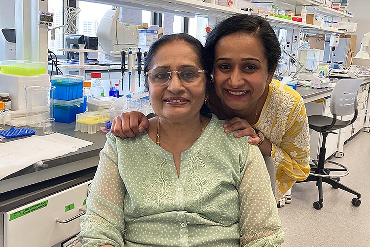 Two women from India look at the camera and smile. The younger one is standing behind the older one, with her hands on the older's shoulders.