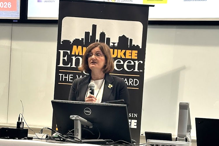 A woman speaking at a podium. She has brown hair and is wearing a dark suit jacket. A college banner is behind her. A woman speaking at a podium. She has brown hair and is wearing a dark suit jacket. A college banner is behind her.