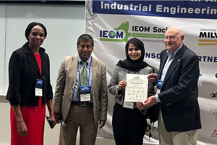 A group shot of four - two women and two men displaying a plaque. One woman is wearing a red dress and the other has a headscarf. One man is dark-skinned and wearing a tan suit. The other is white in a dark suit jacket. A group shot of four - two women and two men displaying a plaque. One woman is wearing a red dress and the other has a headscarf. One man is dark-skinned and wearing a tan suit. The other is white in a dark suit jacket.