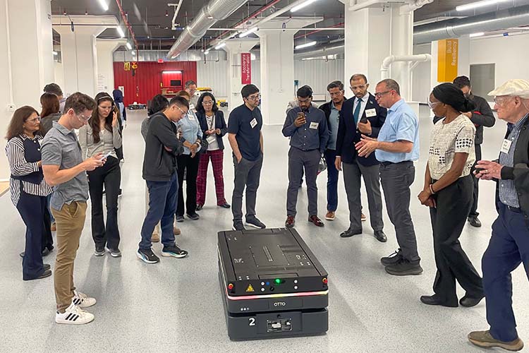 A group of people gather around a trunk-like device in an industrial setting. Most are looking at one person wearing a light blue shirt who is describing the device.