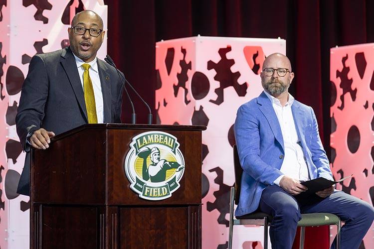 Two men, one in a dark suit speaking at a podium , and another in a light blue jacket sitting on the right. Two men, one in a dark suit speaking at a podium , and another in a light blue jacket sitting on the right.