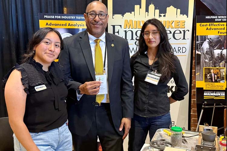 Two women stand on either side of a bald man in glasses and a yellow tie, all looking at the camera with a UWM poster behind them. Two women stand on either side of a bald man in glasses and a yellow tie, all looking at the camera with a UWM poster behind them.