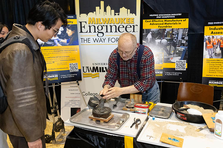 An older man at a table bends over to pour molton tin into a mold to show an onlooker in a brown jacket. An older man at a table bends over to pour molton tin into a mold to show an onlooker in a brown jacket.
