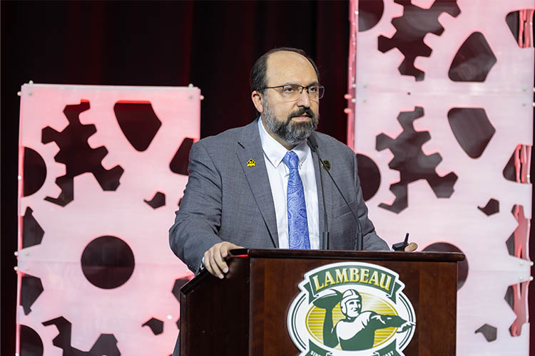 a man with glasses speaks at a podium branded "Lambeau Field." The stage background show pink gear shapes on a black background. a man with glasses speaks at a podium branded "Lambeau Field." The stage background show pink gear shapes on a black background.