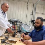 A man standing in a white lab coat on the left assists a man in a blue shirt on the right, seated, as they work on equipment at a table