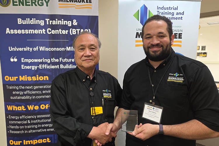 Two men are shaking hands while they look at the camera. They are both wearing black and are standing in front of an ITAC poster.