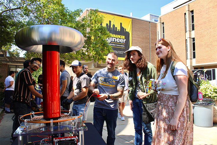A male instructor demonstrates a Tesla coil to students gathered outdoors in front of a Milwaukee Engineer sign.