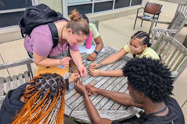 A female college student wearing a backpack leans over a table with four high school students - all African American - seated with arms outstretched on the tabletop.