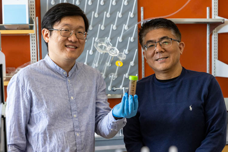 Two men standing next to one another with the one on the left wearing a light blue shirt and holding a water sample in a tube. The other man is wearing a navy blue shirt.