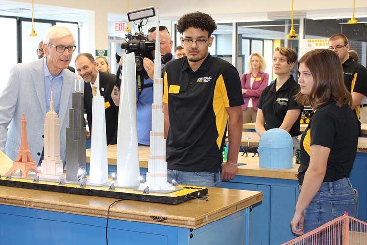 A man with white hair in a light blue jacket visit with college students in back of a model display.