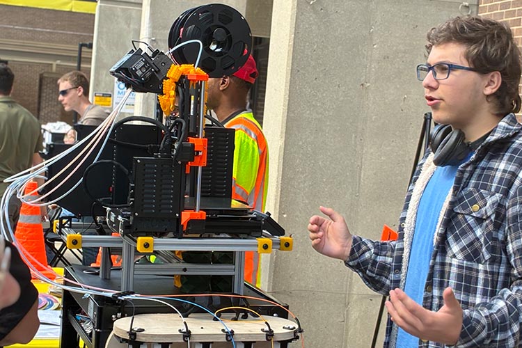 A young many in a blue shirt speaks, standing next to a 3D printer. A young many in a blue shirt speaks, standing next to a 3D printer.