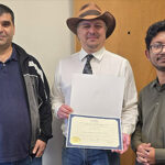 Three men standing in a row looking at the camera. The man in the middle is wearing a hat and holding a certificate.