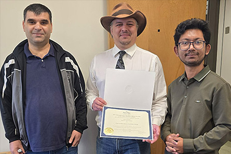 Three men standing in a row looking at the camera. The man in the middle is wearing a hat and holding a certificate.